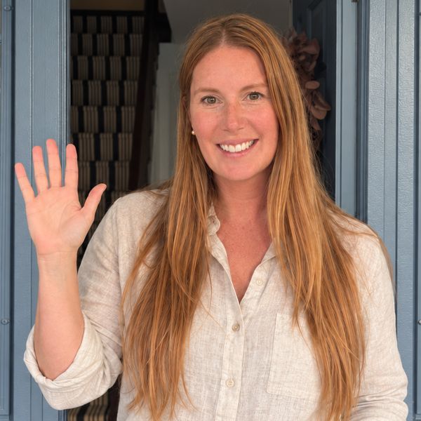 Susie at the open blue front door of her farmhouse in a linen shirt, smiling and waving.